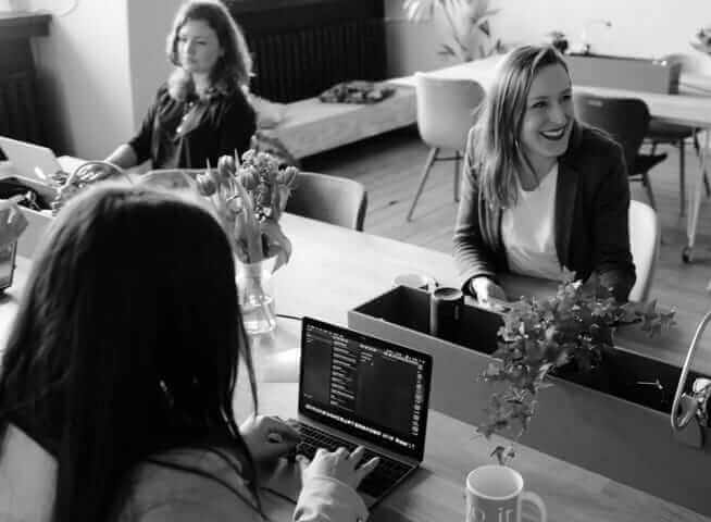 A group of women working together in a modern office, one smiling while others are focused on their laptops.